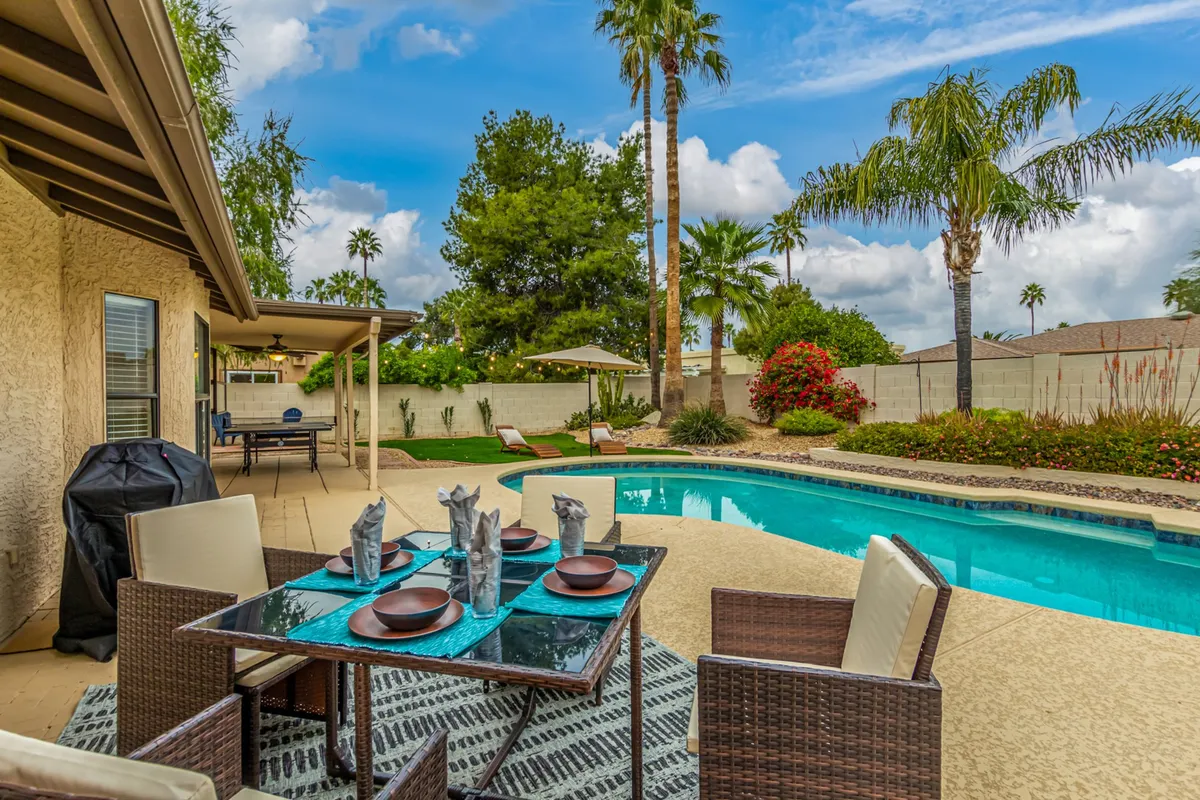 A peaceful yet vibrant backyard featuring a pool, outdoor dining table, palm trees, and colorful flowers. The setup invites both daytime poolside relaxation and memorable starlit evenings. The scene radiates enjoyment and togetherness in a picturesque Arizona setting.