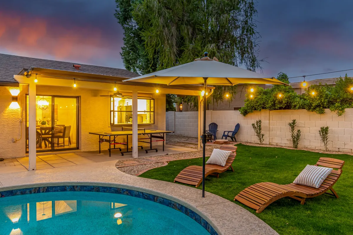 A vibrant backyard scene at sunset featuring a sparkling swimming pool, lounge chairs, a table tennis table placed on the patio, and string lights overhead. The patio area invites relaxation while the spacious pool area is ready for swimming and group games.