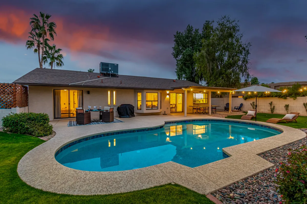 An inviting exterior view that showcases the swimming pool with lounge chairs and a spacious outdoor dining area. The landscaped garden frames the social hub, with string lights creating a magical evening ambiance. The setting is designed for both relaxation and lively meals, echoing the theme of al fresco dining under the desert sky.