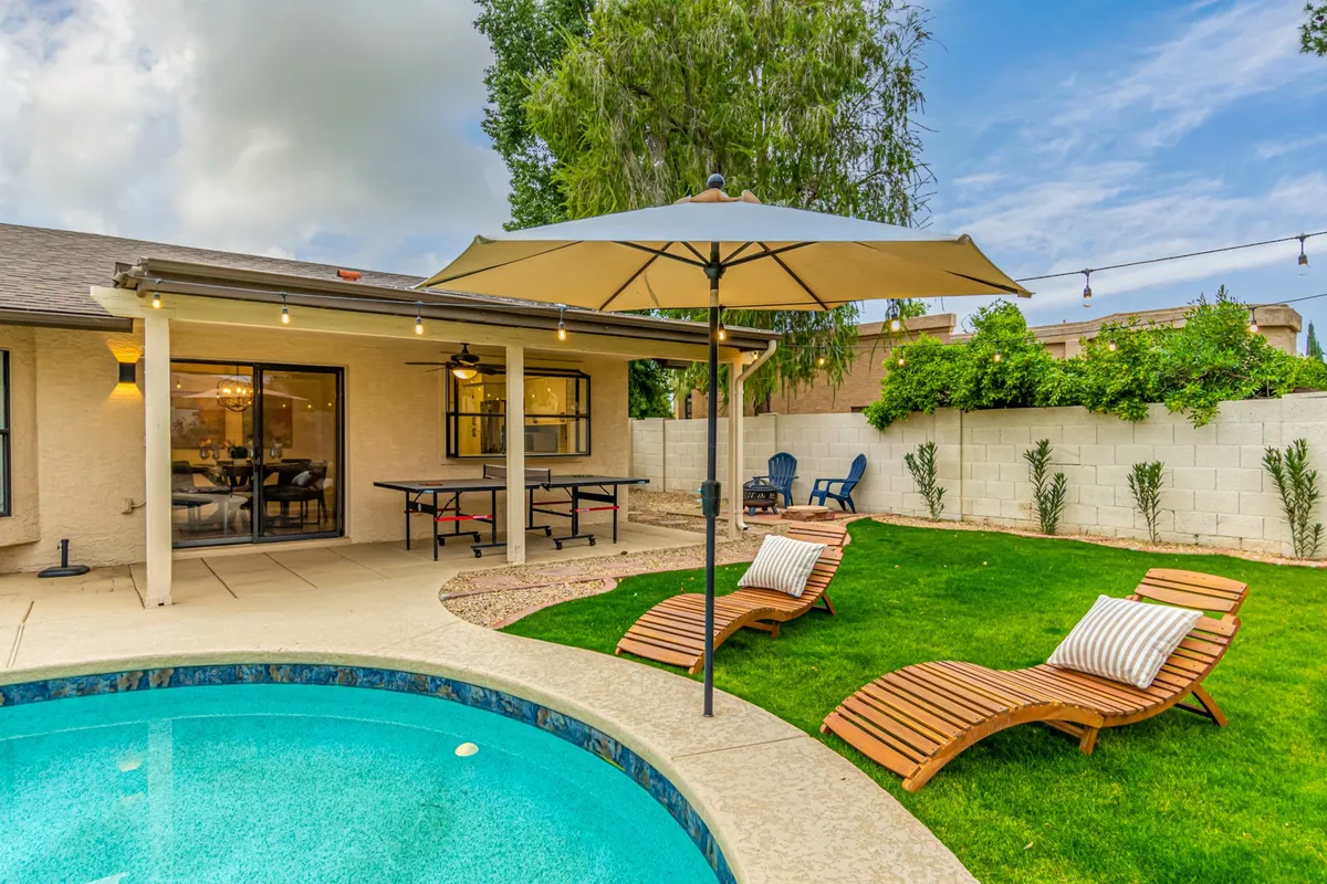 A tranquil outdoor space with a shimmering swimming pool, several lounge chairs, a dining table shaded by a patio umbrella, manicured green grass, and decorative plants. The vibe is serene and leisurely, ideal for both quiet mornings and lively afternoon gatherings.