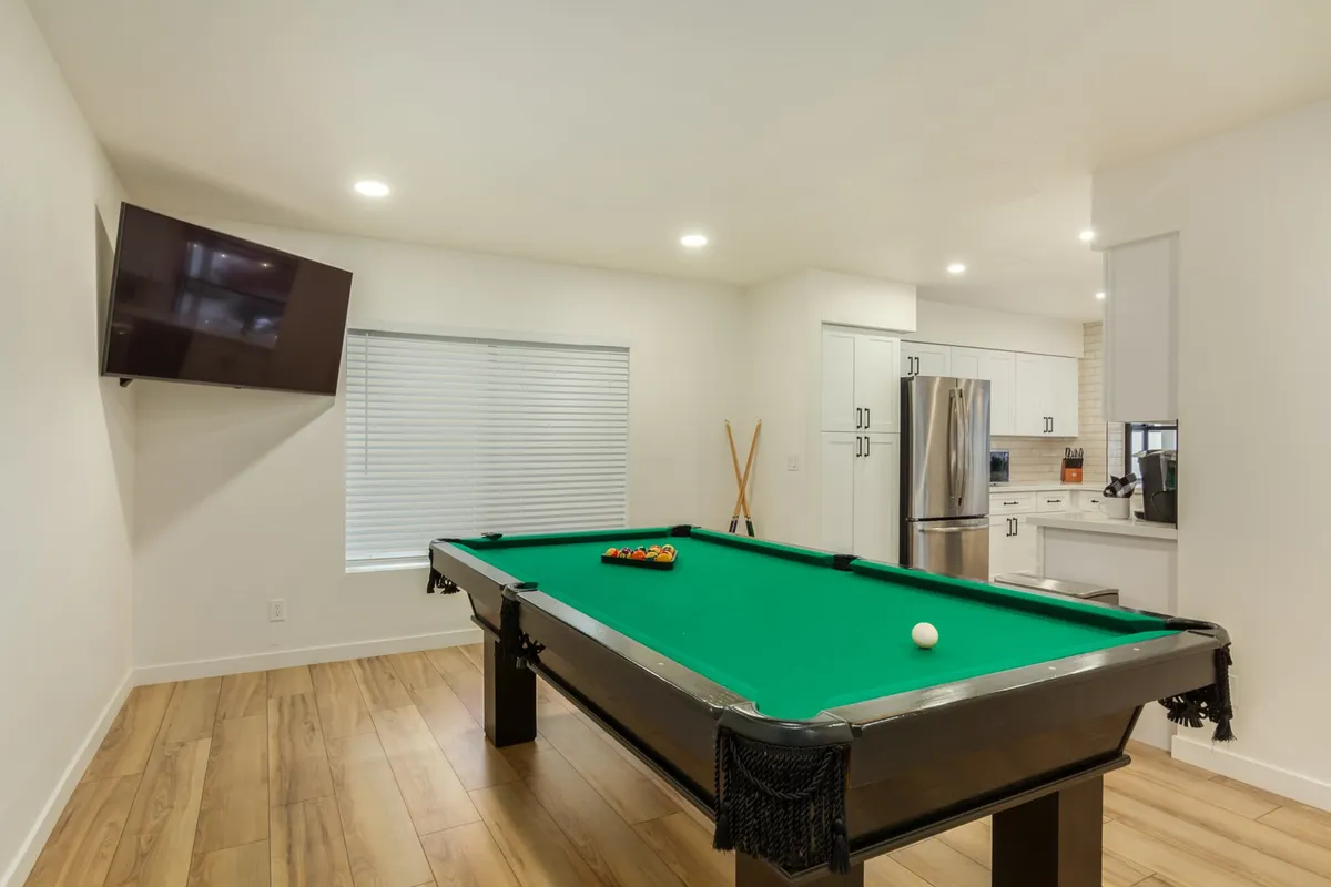 This image captures a vibrant game room featuring a pool table as its centerpiece, a large television mounted on the wall, and an adjacent kitchen area visible through the open plan. Natural light streams in from a window with blinds, creating a lively and welcoming atmosphere ideal for socializing and entertainment.