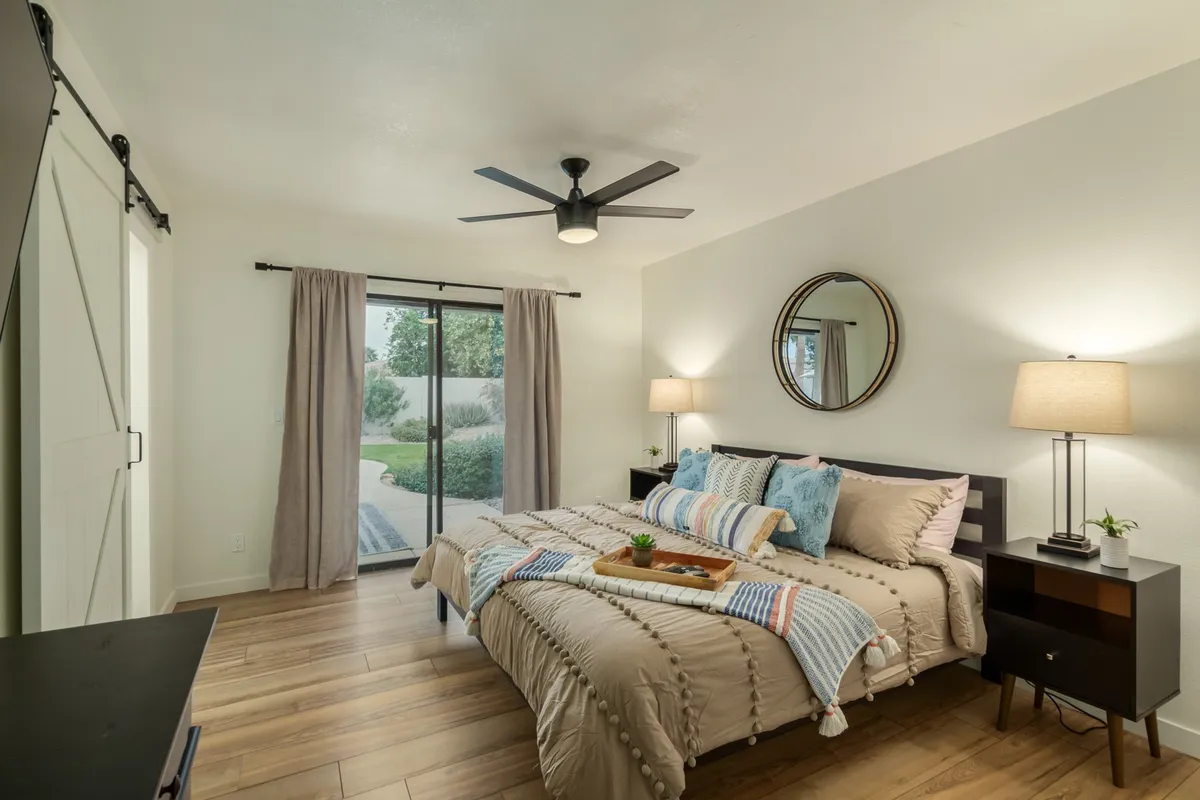 A serene bedroom featuring a neatly made bed with decorative pillows, flanked by nightstands with lamps. The soft light from the curtains and sliding doors gives the space a gentle glow, while a round mirror adds a touch of style and spaciousness.