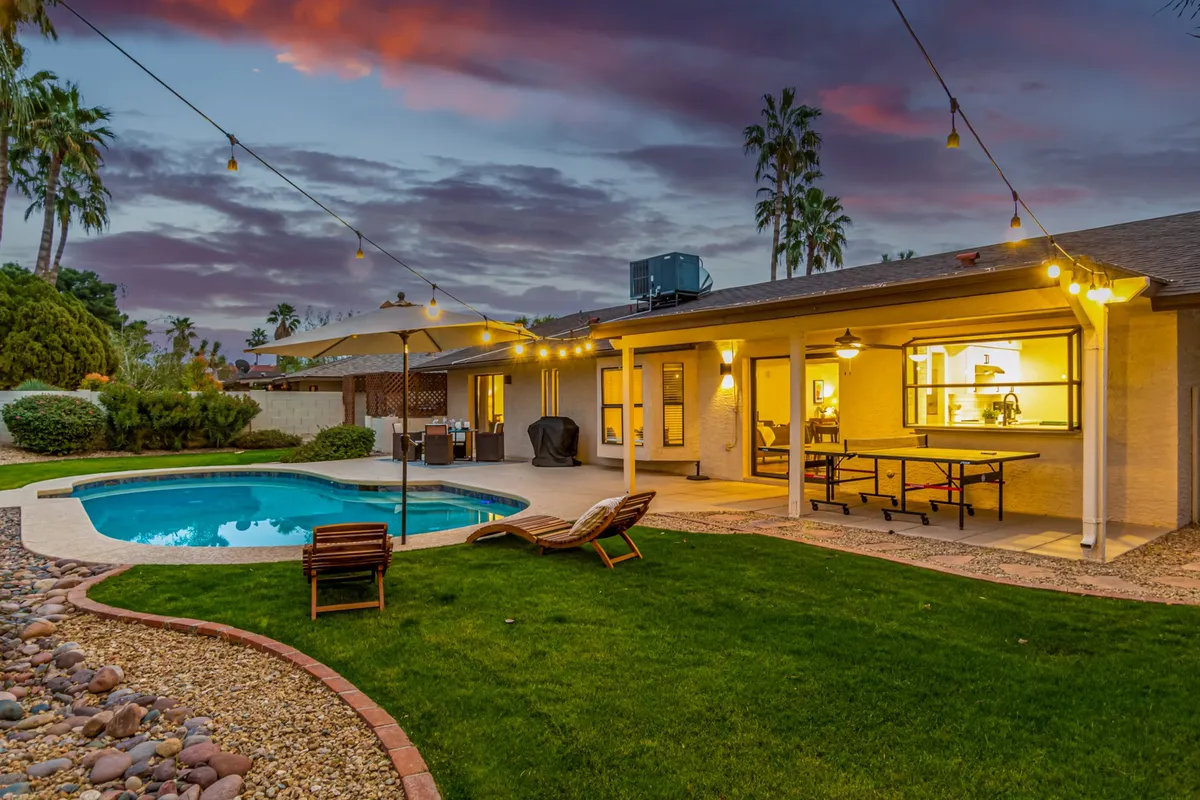 A sun-kissed backyard retreat featuring a sparkling swimming pool, inviting outdoor seating area, string lights overhead, a dining table, and lush greenery all around. The atmosphere is casual, open, and perfect for unwinding after a day of golf or hosting a relaxed evening gathering.