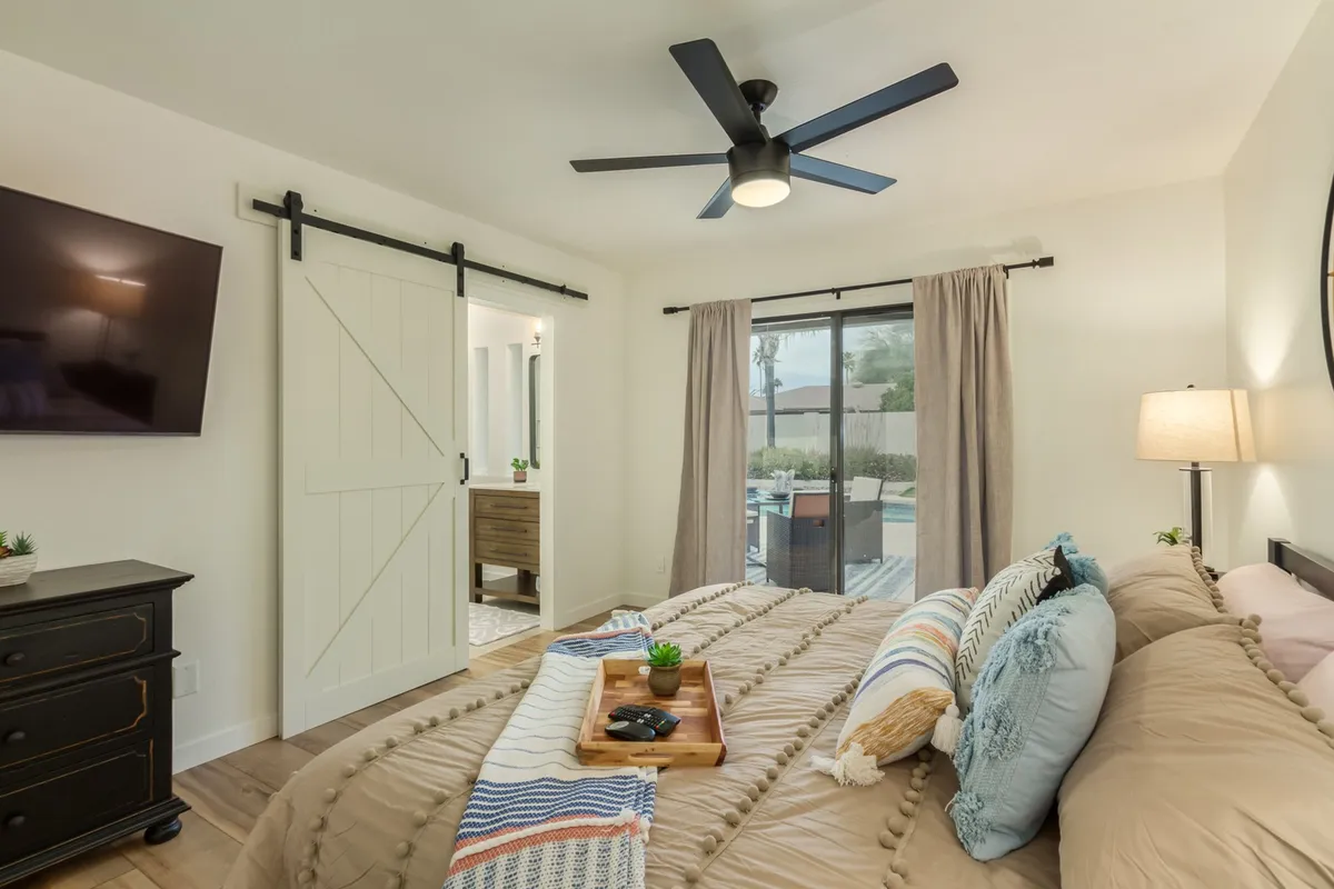 A tranquil bedroom with a comfortable bed, decorative pillows, a sliding barn door, large window with curtains, ceiling fan, and a television. The room is filled with natural light, creating an inviting and restful environment.