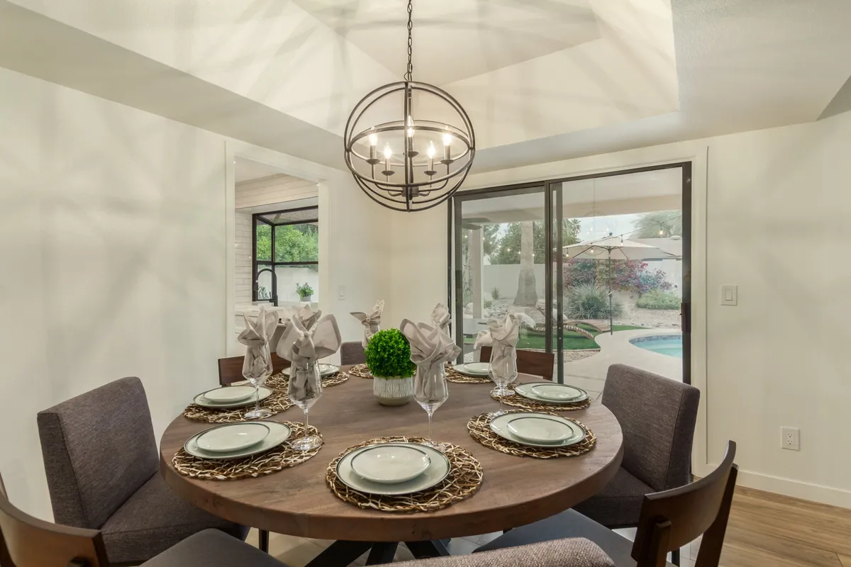 A round wooden dining table elegantly set for eight, centered beneath a striking chandelier, sits ready for memorable meals and engaging conversations. A green centerpiece and neat place settings add an inviting, celebratory touch.