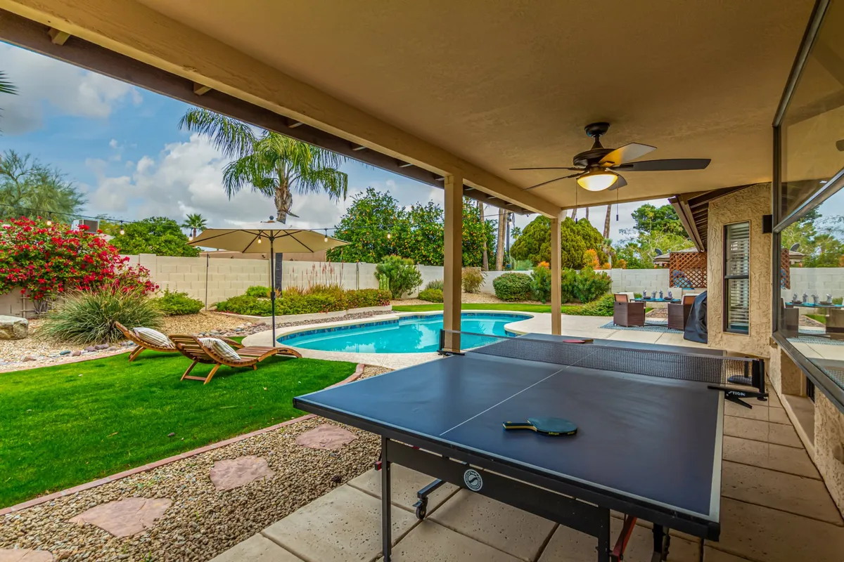 A relaxed outdoor area with a ping pong table beside the swimming pool, surrounded by lounge chairs and greenery. A large outdoor umbrella provides shade, suggesting enjoyable days spent outside, whether playing games or relaxing poolside.