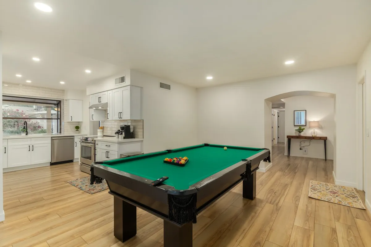 A relaxed interior scene showing the pool table prominently situated between an open kitchen area and a cozy social space. An arched doorway adds architectural interest, while the setup invites friendly competition and togetherness, blending the culinary and entertainment zones seamlessly.