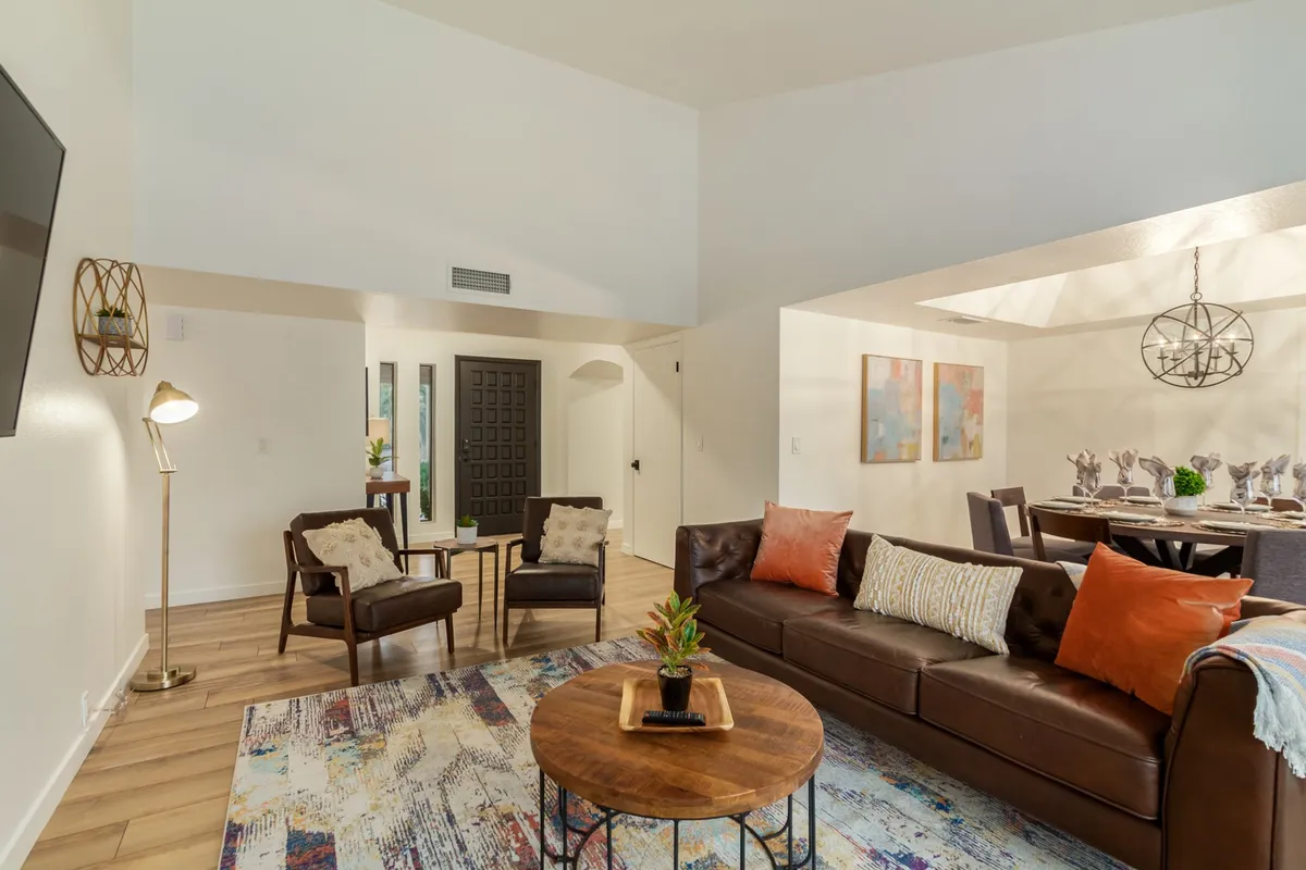This image captures a warm, open-concept living and dining space. It features a leather sofa, a round wooden coffee table, accent chairs, and a dining area with an elegant chandelier in the background. Large windows allow natural light to flood the space, highlighting the contemporary décor and art on the walls.