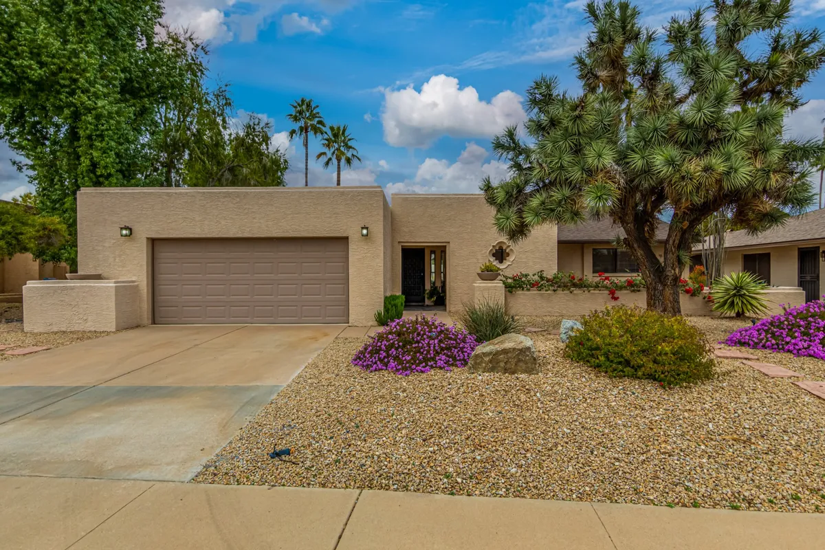 A view of the landscaped front yard shows meticulously maintained flower beds, native cacti, and a stone pathway leading to the home. The broad front yard is basked in sunlight, with inviting spaces for outdoor relaxation and potential for morning or evening strolls.